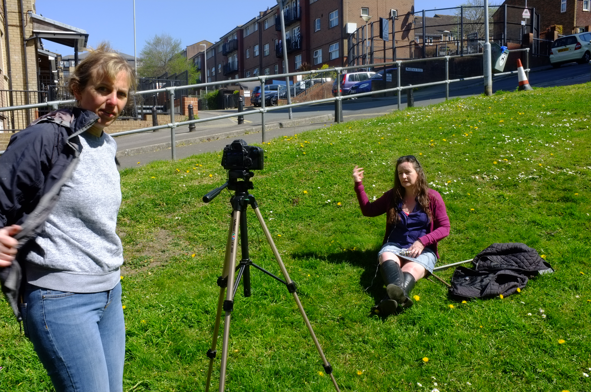 A photo showing a woman standing by a camera and another woman sat on the grass behind, waving.