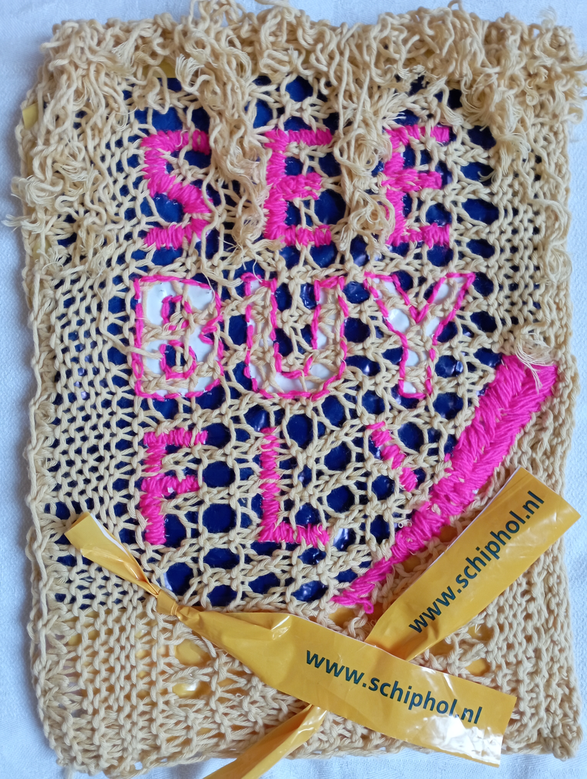 "See, Buy, Fly. The Web of Life is Torn by Consumerism."  Materials: netting knitted with essentials Organic Cotton by Rico, mustard colorway, color 003; plastic bag from Schiphol Airport Duty Free Shop, and 100% acrylic yarn from ON line Linie 50, Prima, color 0033 used for embroidery