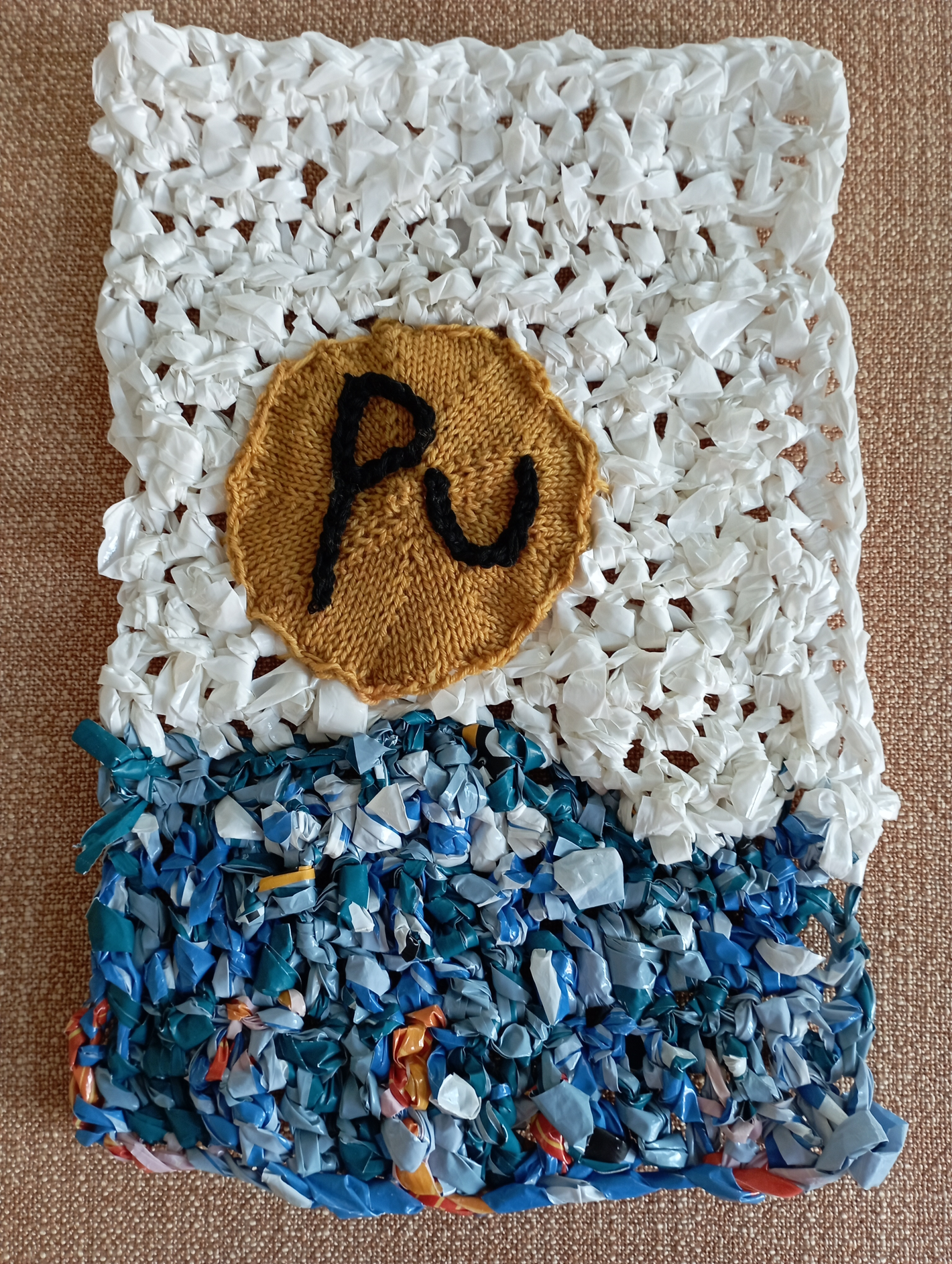 "Plastic Pollution in the Oceans has Reached Critical Mass" , the latter symbolized by the sphere of Plutonium, approx the size of the critical mass for a nuclear bomb.  Materials: Plastic Yarn (Plarn) for background (crochet), sphere knit from hand-dyed Merino Sock woll (scrap) and black embriodery stitched on