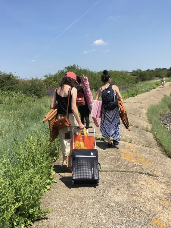 Three figures photographed from behind walking along a concrete path near a green verge heavily laden with a suitcase and other equipment. The sky is very blue.