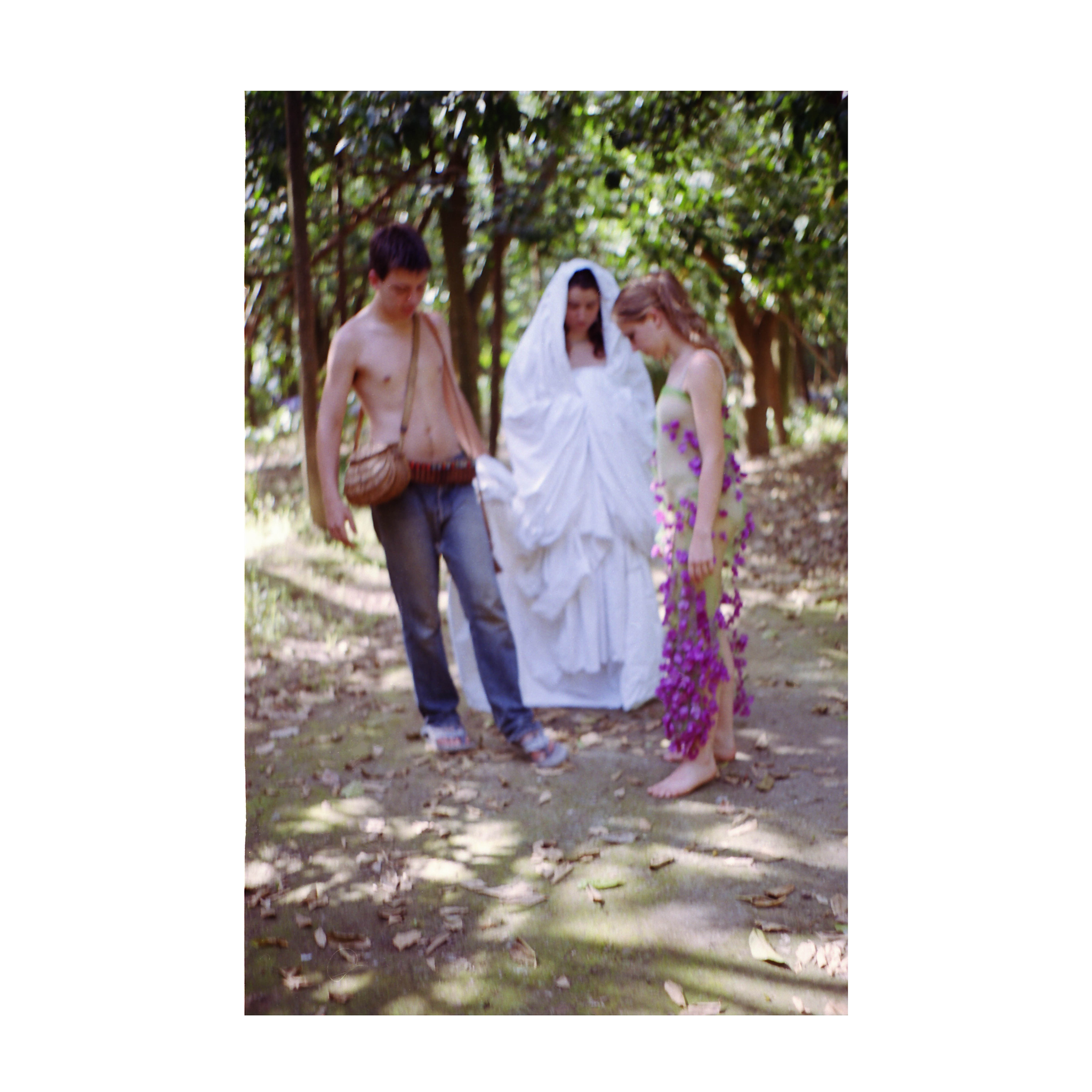 The photograph shows three figures standing on a leaf-scattered path in a wooded area. On the left, a shirtless person wears jeans and a basket bag slung across their shoulder. In the center, a person is draped in layered white fabric that covers their body and head. On the right, another person wears a light-colored transparent dress covered with purple flowers and stands barefoot, looking down. The background is filled with trees and dappled light.