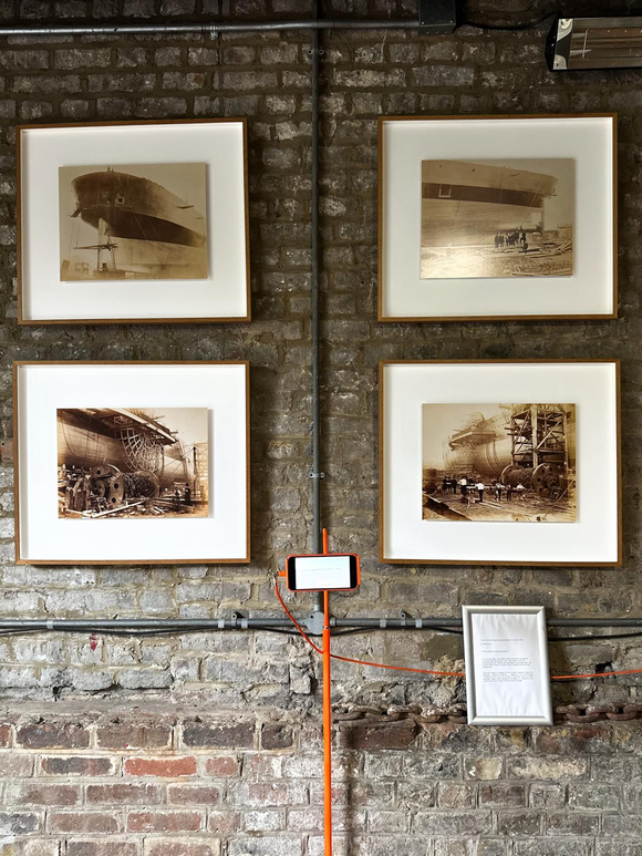 image of orange mobile phone on a stand in front of historic framed black and white prints of old photos of industry related to the Brunels