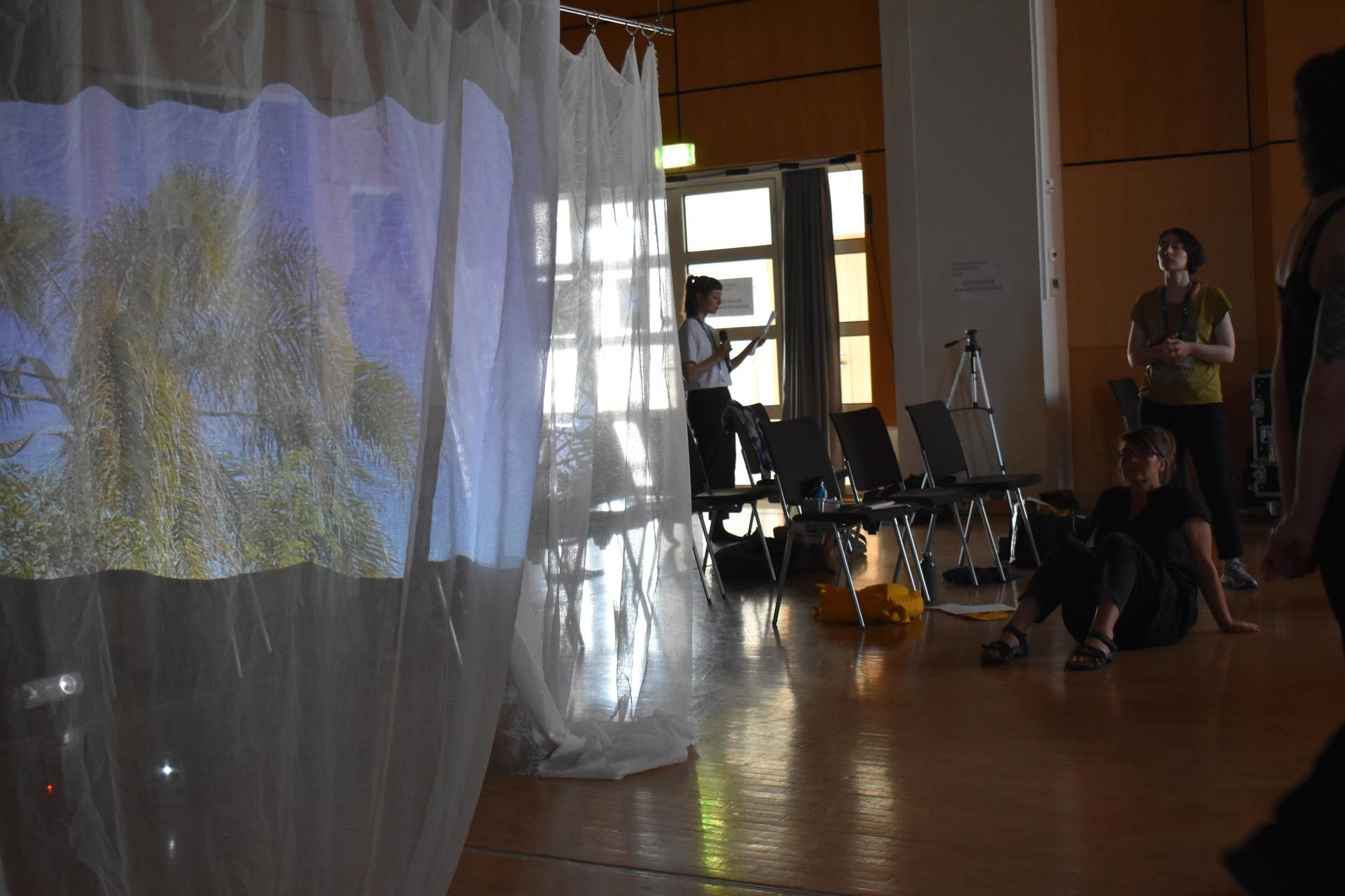 A projection of palm trees with blue sky is projected onto the semi-opaque curtain. There are empty chairs from the conference and people stand or sit listening to the work.