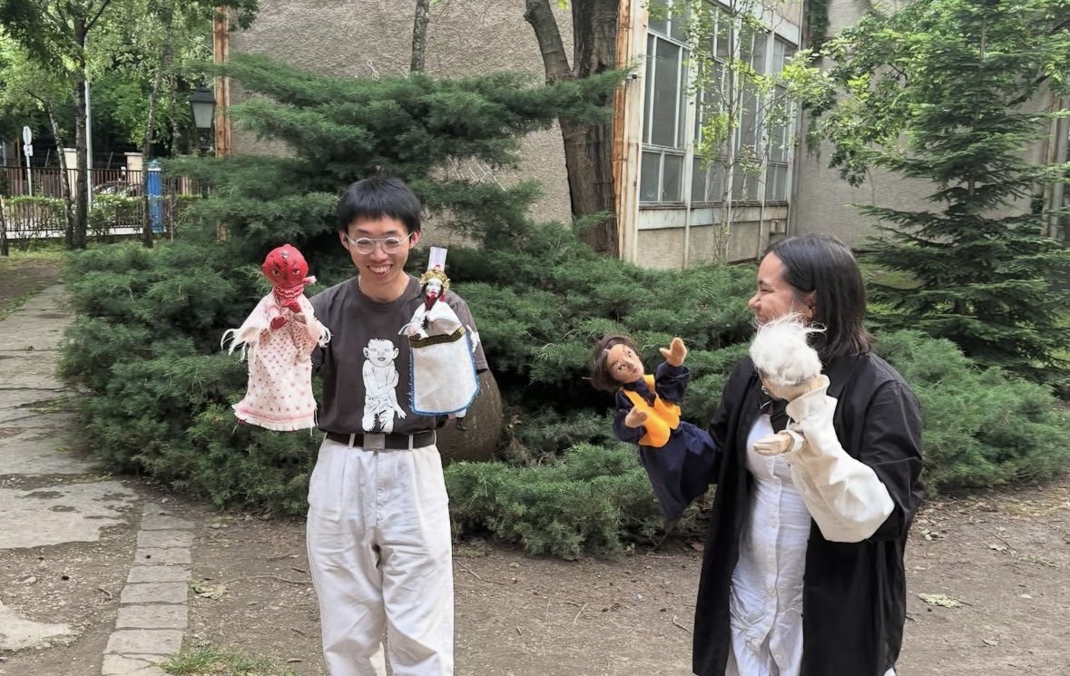 Two people standing in front of a building, bushes and trees. Both hold a puppet in each hand and smile.