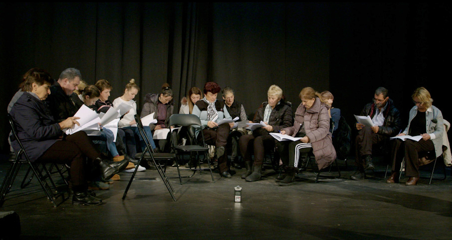 A group of 16 people sitting in a semicircle on a black stage with black curtains behind them. They are collectively reading The Chimneys on the Adriatic. A recording device is placed in the middle on the floor.