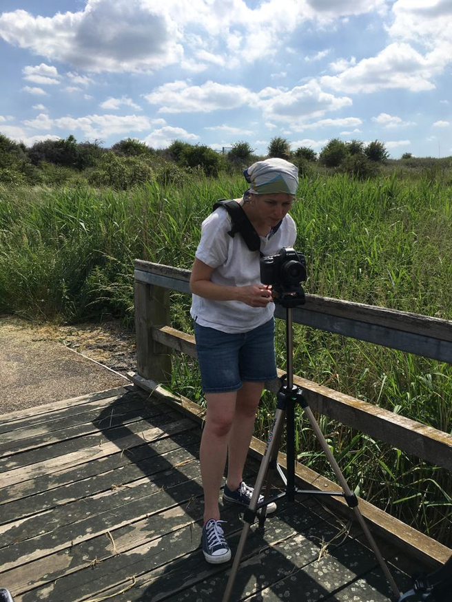 A white woman in a white t shirt stands behind a DSLR on a tripod looking at the display. She is on a wooden bridge with green reeds behind her and the sky is blue with lots of small white/grey clouds.
