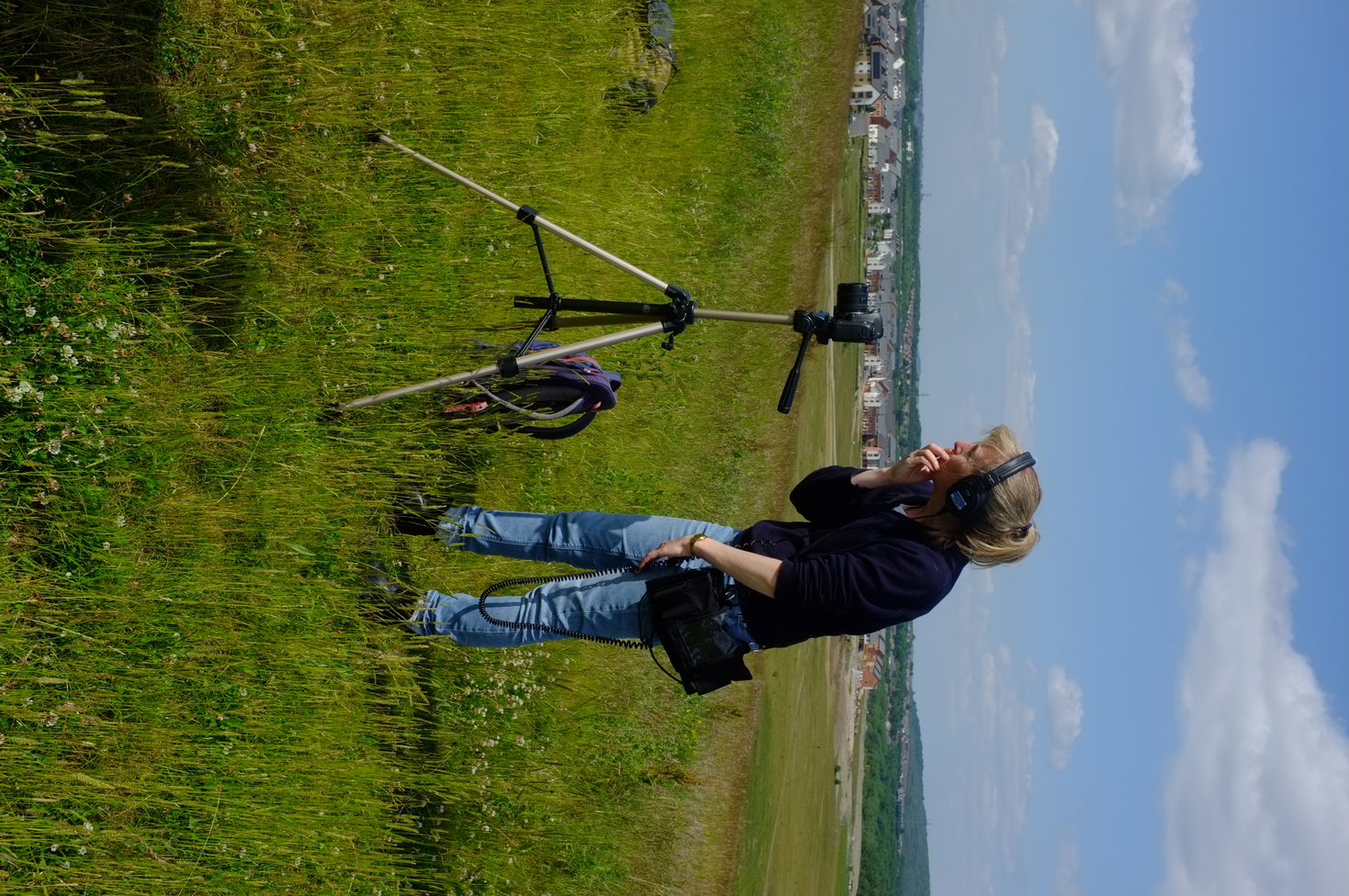 A white woman wearing blue and navy with headphones on  looks on as she films using a camera on a tripod