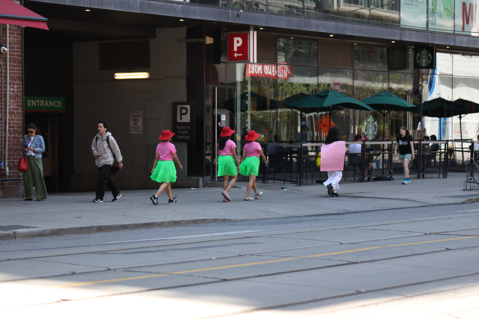 A walk with a playful question, 'To cake or not to cake?', invited pedestrians and patio patrons at the popular destination of King and Portland to reflect within a space largely defined by consumerism, in response to Score 4 created by Mushtari Afroz.
The walk was led by Mushtari, with the question displayed on a piece of pink art paper hanging from her back as she strolled along the sidewalk, carrying a piece of cake on a tray in her hand.