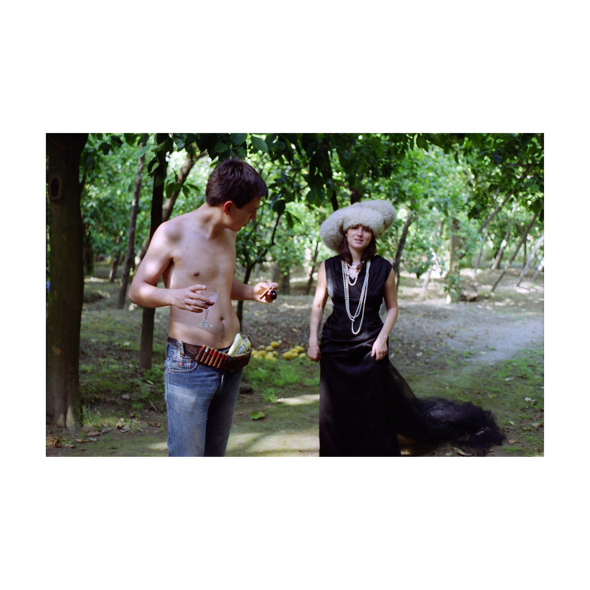 A shirtless young man stands outdoors in a sunlit grove, holding a glass of wine and a small object, with bullets tucked into his jeans’ waistband. Opposite him, a woman dressed in a long black gown, layered pearl necklaces, and a large fur hat looks toward the camera. The background shows trees and dappled light filtering through the foliage