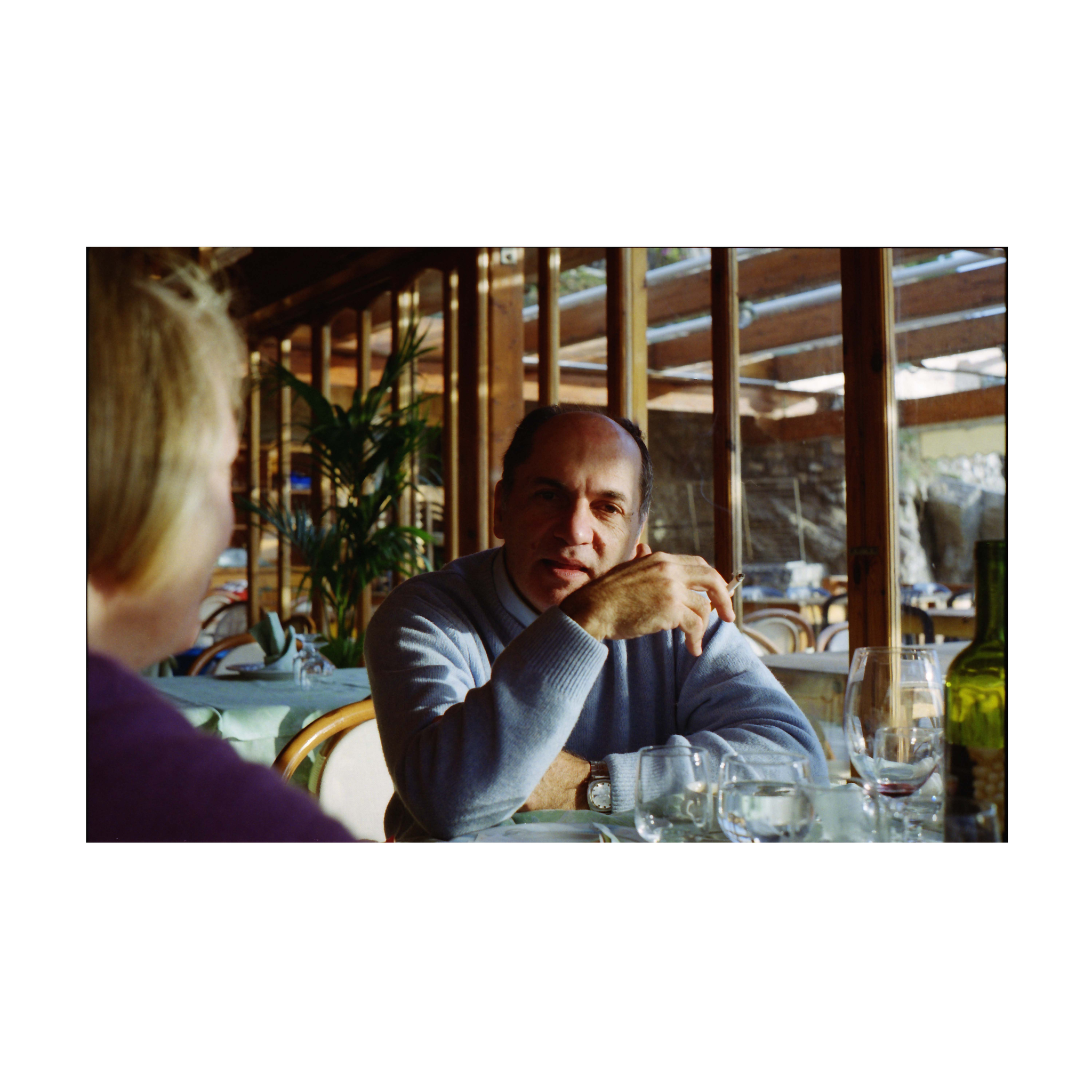 A man is seated at a restaurant table, looking toward the camera. He is wearing a light blue sweater and holding a cigarette in his right hand. In the foreground, the back of another person’s head and shoulder are visible. On the table, there are empty glasses, a green bottle, and some plates. Behind him, there are wooden-framed windows with reflections of the interior and some plants visible outside.