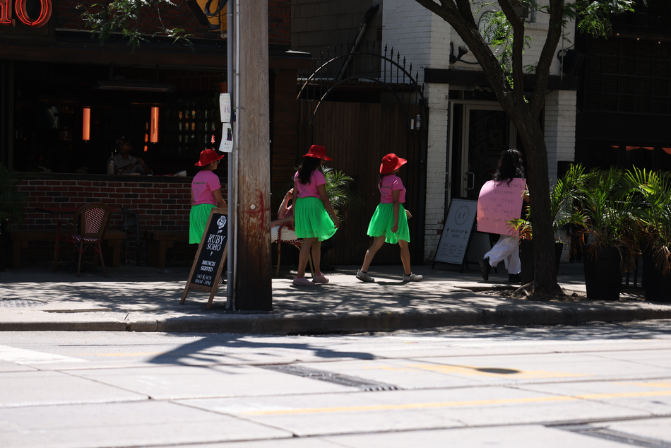 A walk with a playful question, 'To cake or not to cake?', invited pedestrians and patio patrons at the popular destination of King and Portland to reflect within a space largely defined by consumerism, in response to Score 4 created by Mushtari Afroz.
The walk was led by Mushtari, with the question displayed on a piece of pink art paper hanging from her back as she strolled along the sidewalk, carrying a piece of cake on a tray in her hand.
