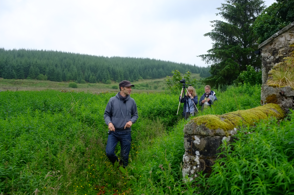 three white figures walking along an overgrown path near a mossy wall. One is carrying a camera on a tripod.