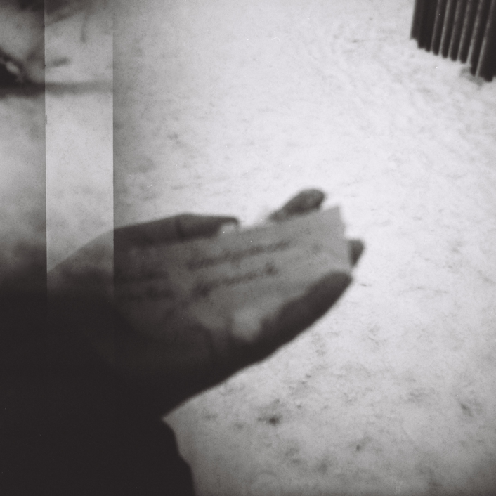 Rakeinen kuva kädestä, joka pitelee pientä paperilappusta, jolla on tekstiä, josta ei kuitenkaan saa selvää. Taustalla näkyy talvinen, valkoinen lumimassa.