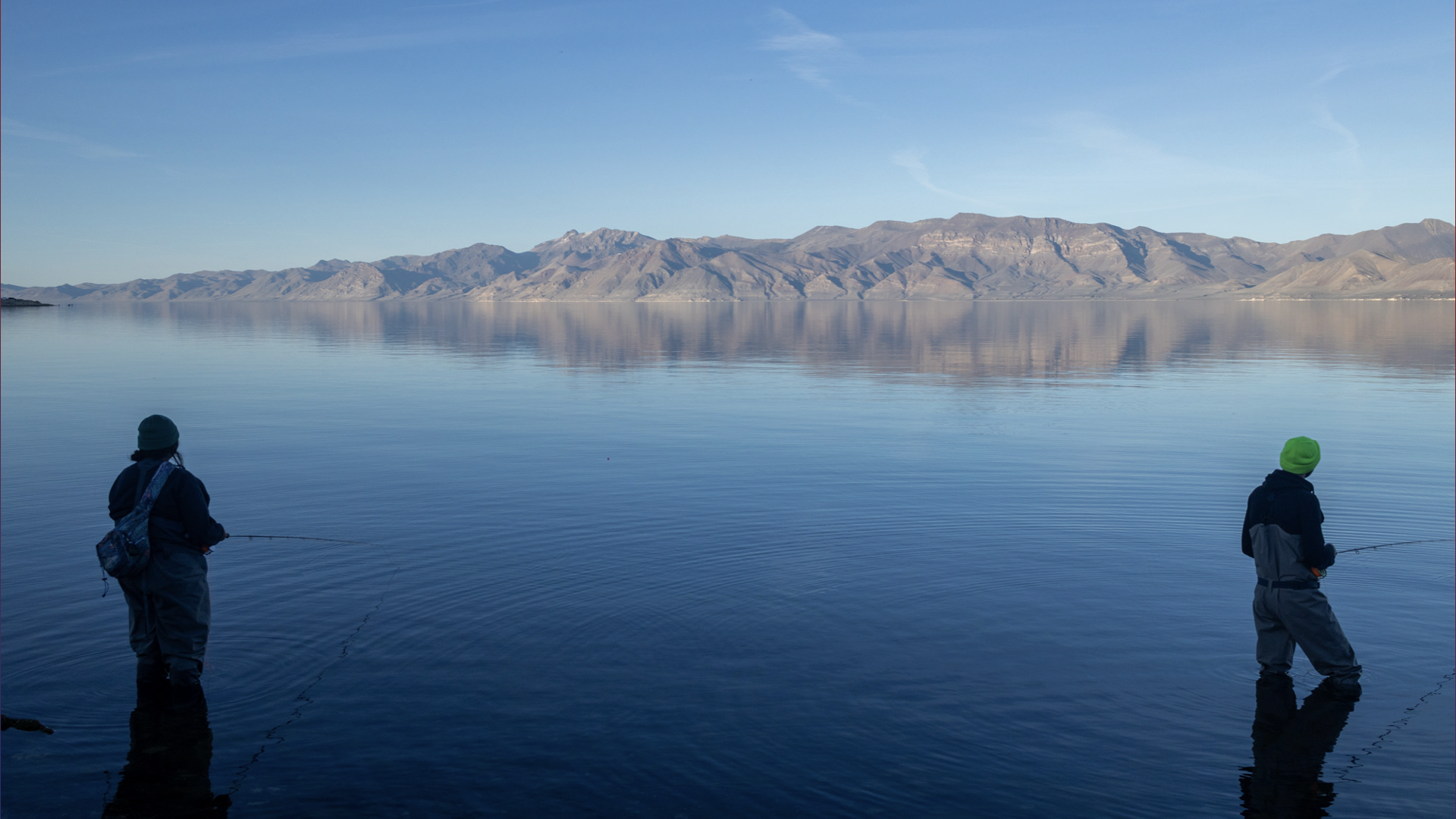 Two fly-fishers viewed from behind standing in the Kooyooe Pa’a lake. Water and mountains on the horizon under a blue sky.