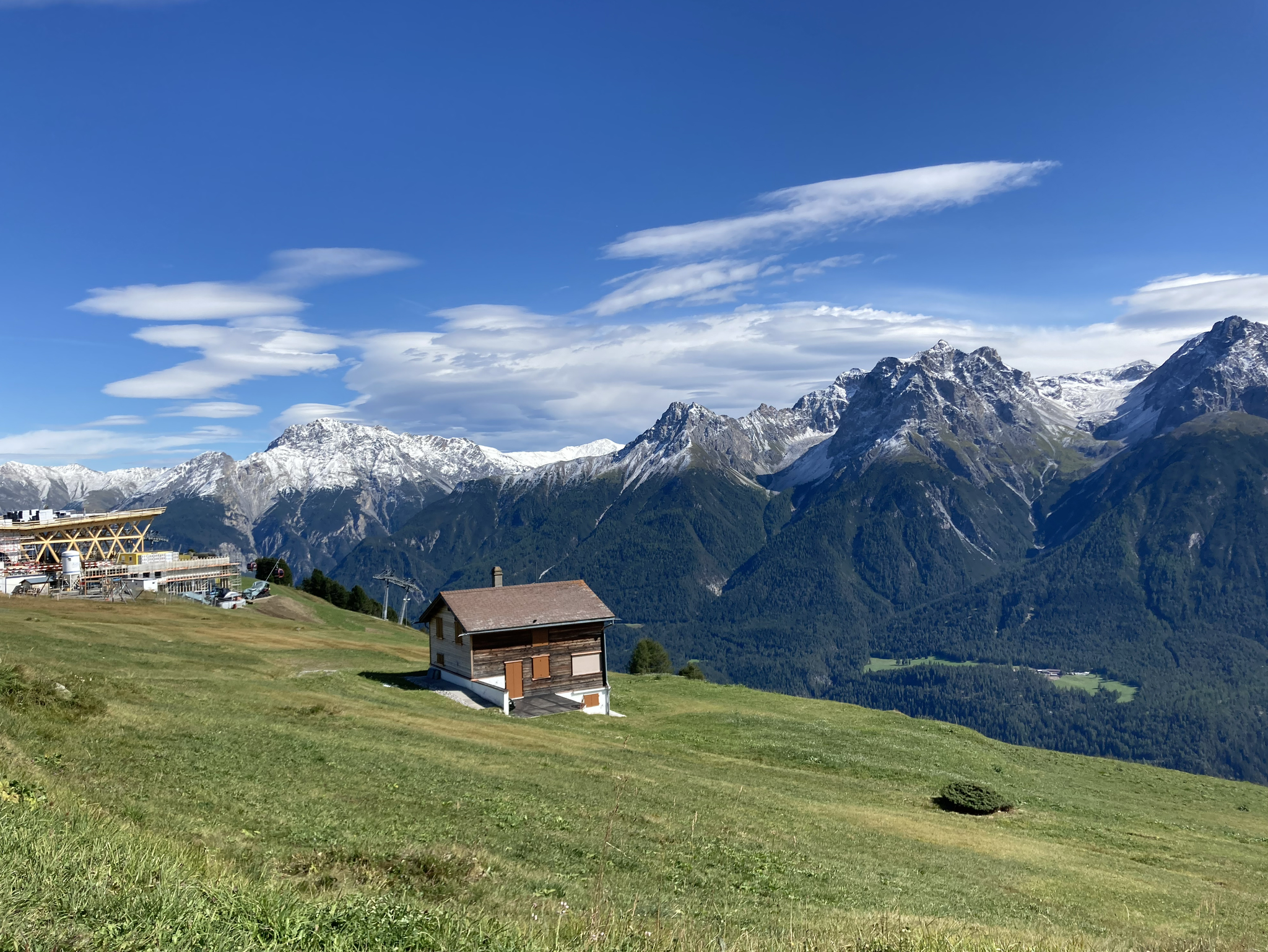 There is a small hut on an alpine pasture. It faces the mountains. To it’s left is a cable car station, still under construction.