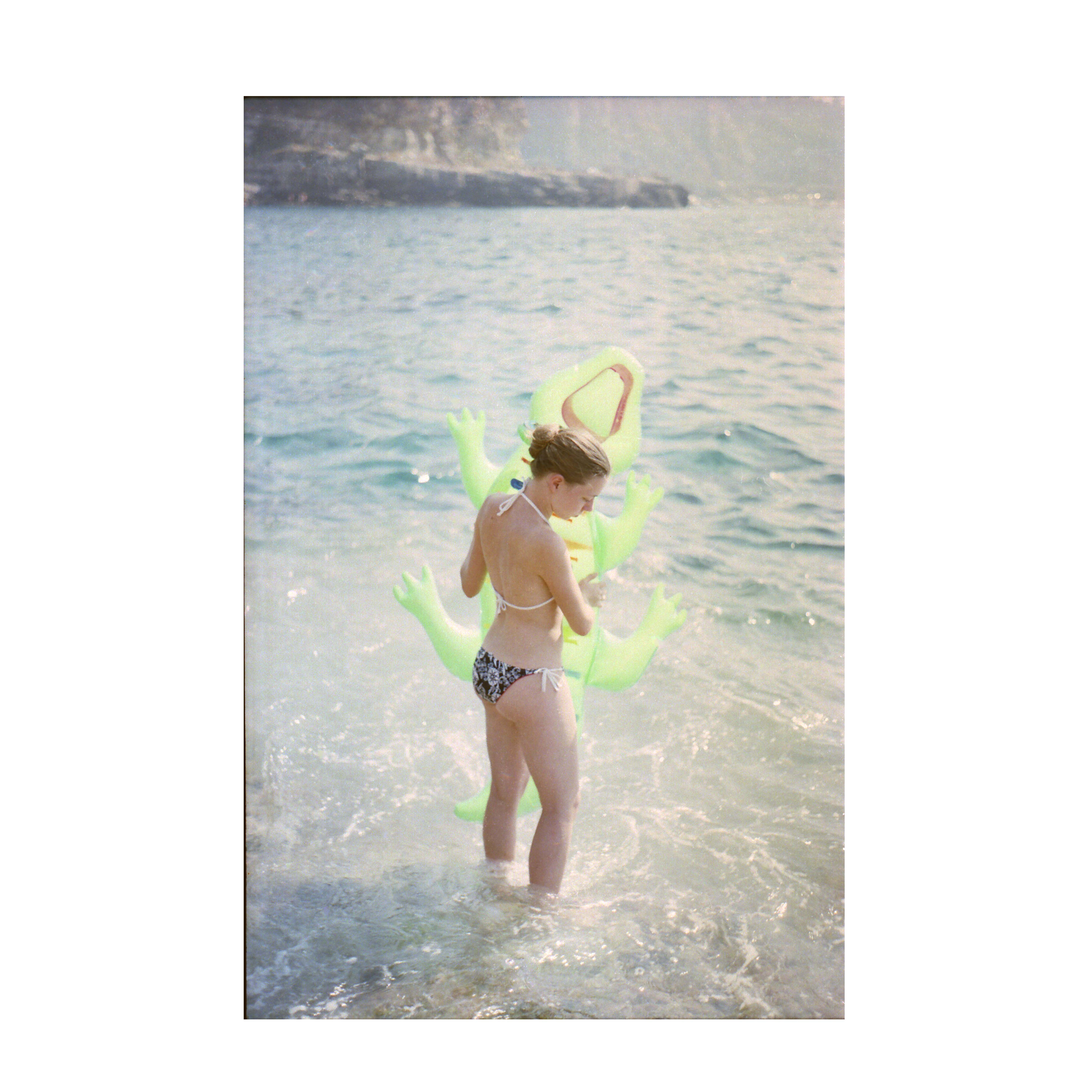 The photograph shows a person standing in shallow seawater, wearing a bikini with a halter-neck top and patterned bottoms. They are holding a large inflatable green crocodile. The sea stretches out into the background with soft waves, and rocky cliffs are visible in the distance under hazy light. The overall tones are pale and sunlit, with the bright green inflatable standing out against the muted blues and greys of the sea and rocks.
