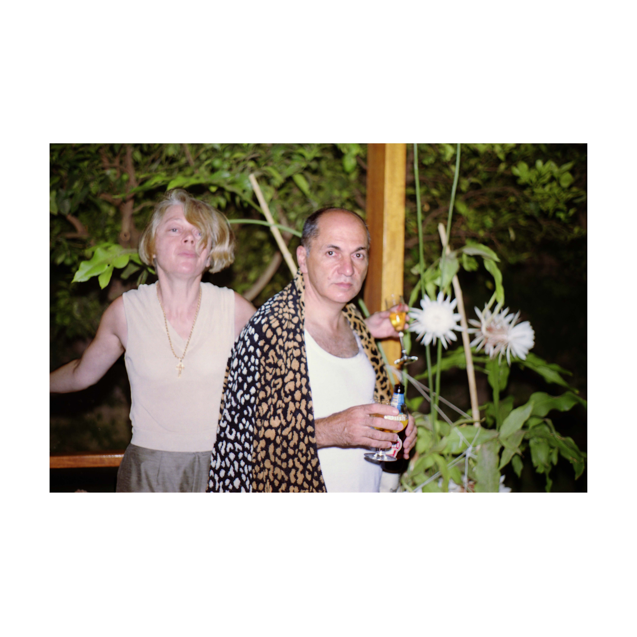 A woman and a man stand outdoors at night beside large white Epiphyllum flowers. The woman, wearing a sleeveless beige top and necklace, is caught mid-movement with her head slightly tilted, her mouth pursed and eyes partly closed, a glass in her hand. The man, wearing a white vest and draped in a patterned shawl, looks directly at the camera with a steady, neutral expression. He holds a drink in one hand and a bottle in the other.