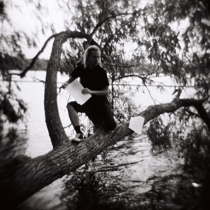 Epätarkka ihmishahmo puussa, veden äärellä, runopaperin kanssa.