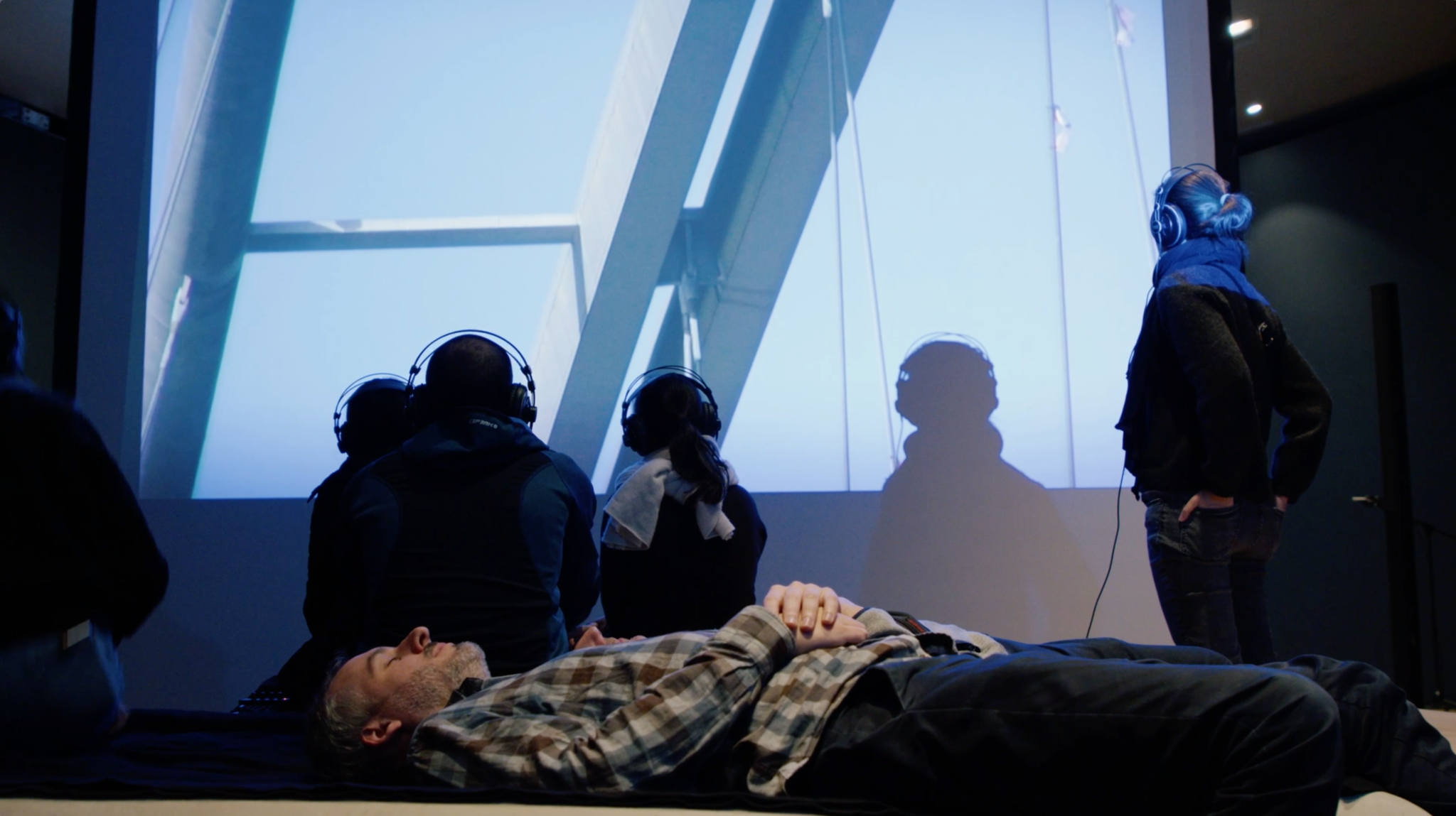 Several people wearing headphones are seated or lying down in a dark room, facing a large projection showing a close-up of a modern architectural structure against the sky. One person lies on the floor in the foreground with eyes closed and hands folded, while others sit upright, absorbed in the audiovisual experience.