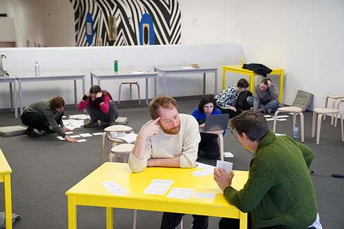 View of the room. Participants sit on tables and on the floor and discuss scenario cards in pairs.