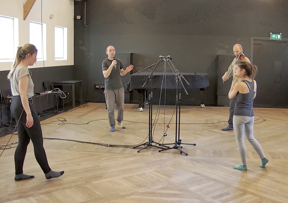 two women, one singing, one playing flute and two men, one singing one playing viola standing around four microphone stands in the middle of a room