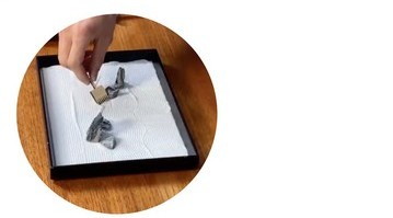 Photo of a hand holding a miniature rake stroking through white sand in a small box.