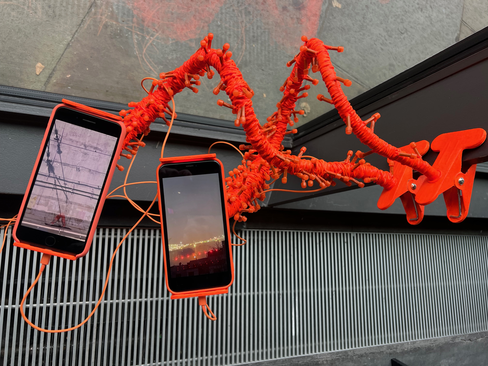 image of artwork installed in gallery. Two hi-vis orange objects holding two iPhones playing two films simultaneously