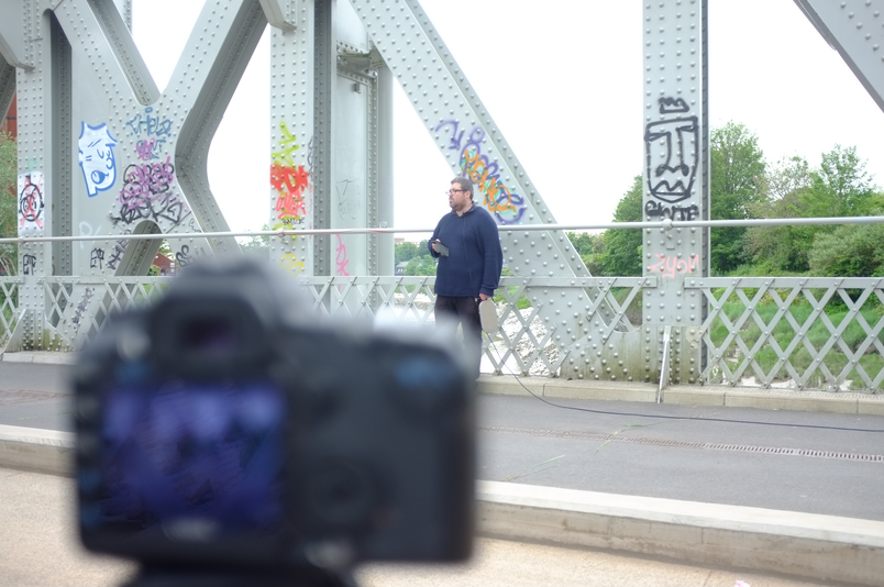 A man dressed in navy stands on wrought iron bridge. In front of him is a DSLR camera, filming the man but the camera and the image on its screen is blurred.