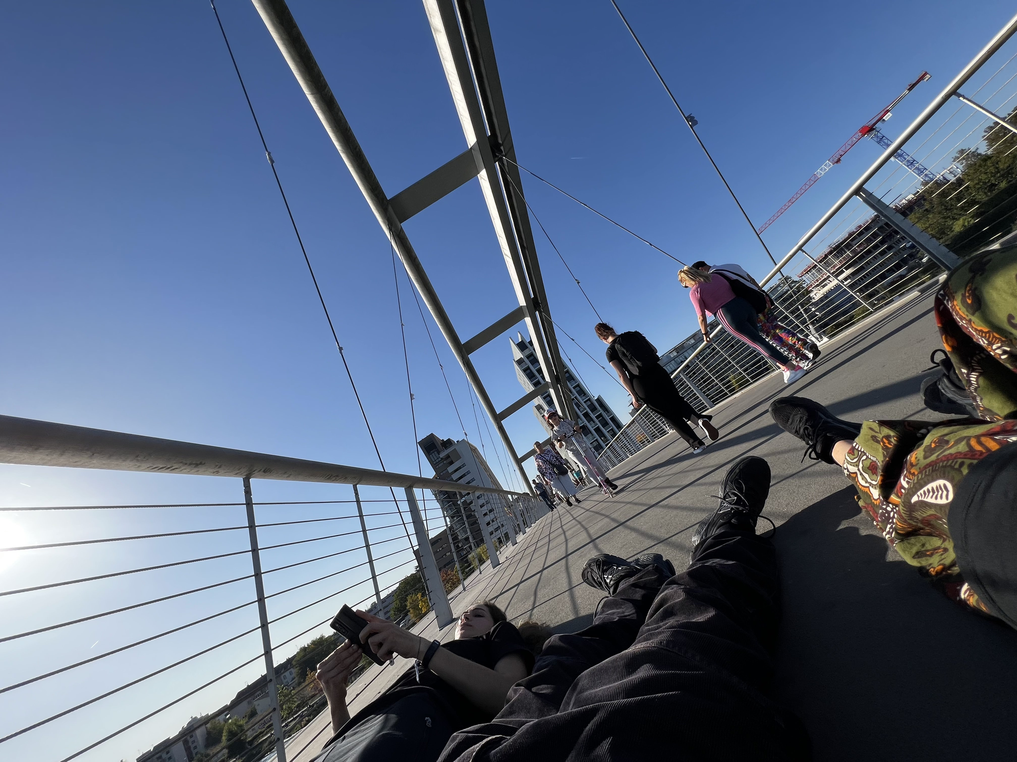 View from the ground of a pedestrian suspension bridge. A group of people lie on the ground in the foreground. Several people walk across the bridge ahead. Clear blue sky with buildings and cranes in the distance.