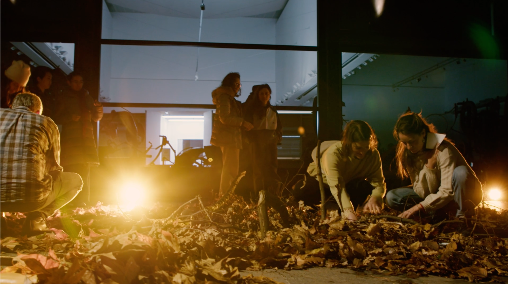 Several people are gathered outside a building at night, illuminated by warm yellow lights. Two people on the right are crouching, arranging dry leaves and branches on the ground, while others stand and observe nearby. The scene suggests a collaborative or playful interaction with natural materials.