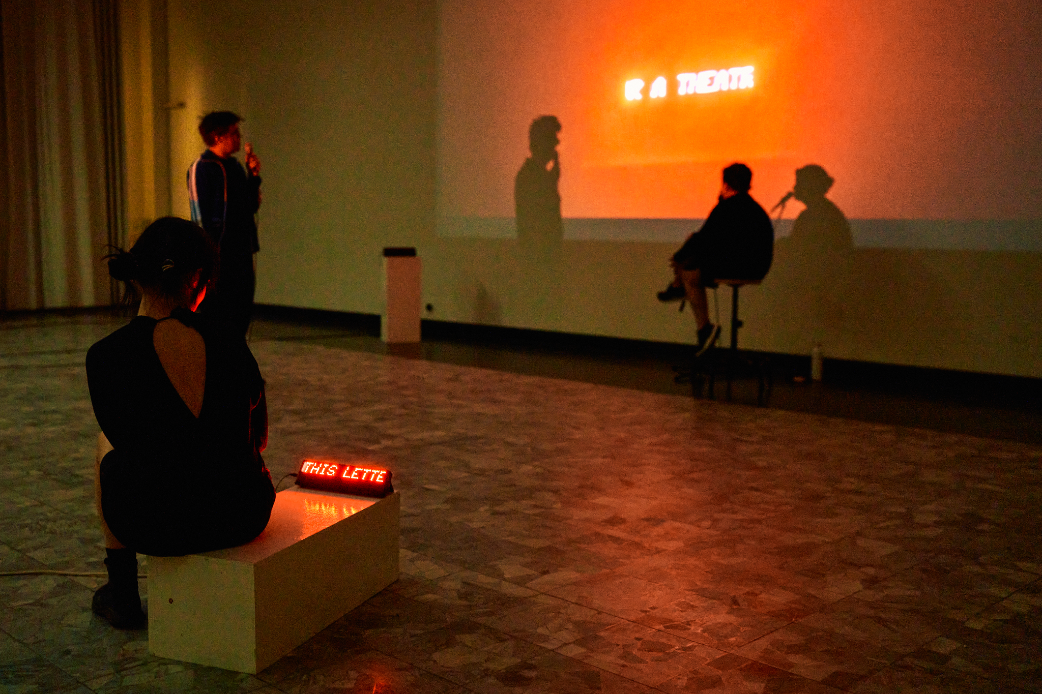Three people in a white box gallery space, one of them watching a running LED ticker placed on a pedestal, and the other two persons watching a video projection that shows a running LED ticker. One of them voices text fragments via a microphone that he holds in his left hand. Photo.