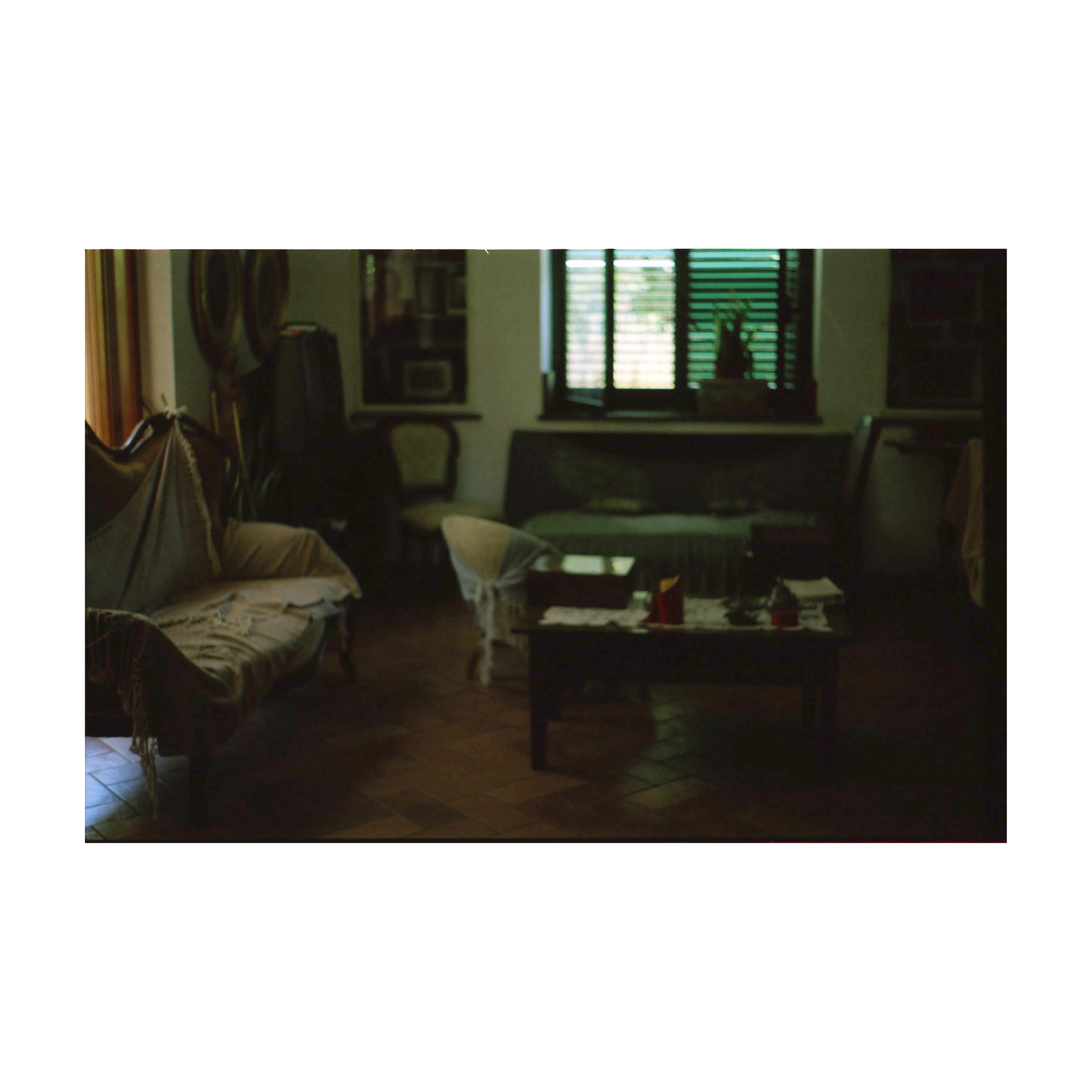 A photograph of a dimly lit living room with vintage furniture. Two sofas are covered with protective cloths, and a coffee table at the center holds scattered papers and small objects. Light filters through a window with green shutters at the back, partially illuminating the space.