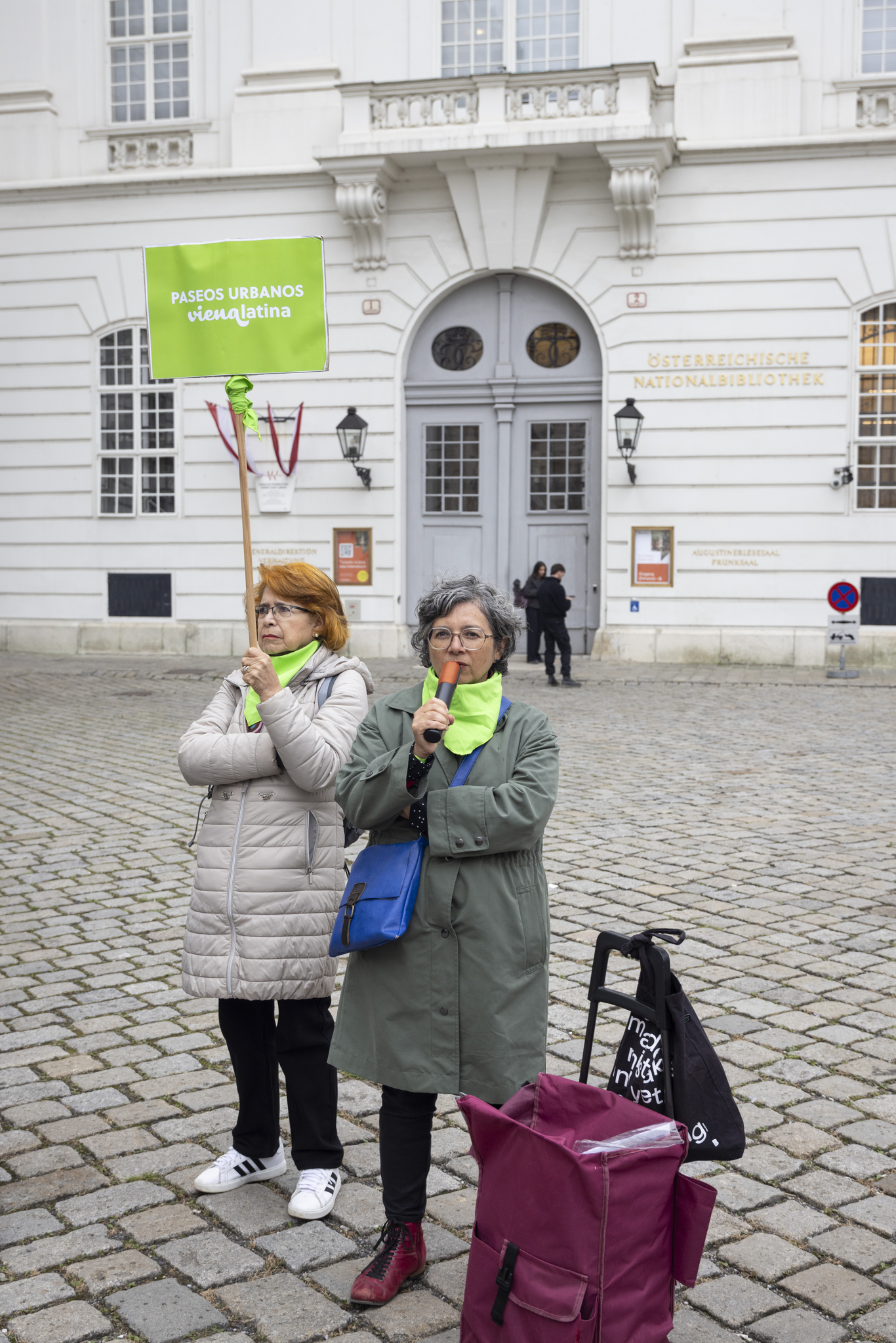Two people standing in front of the National Library in Vienna. One is holding up a sign which says "Paseos Urbanos Viena Latina" and the other is speaking into a microphone.