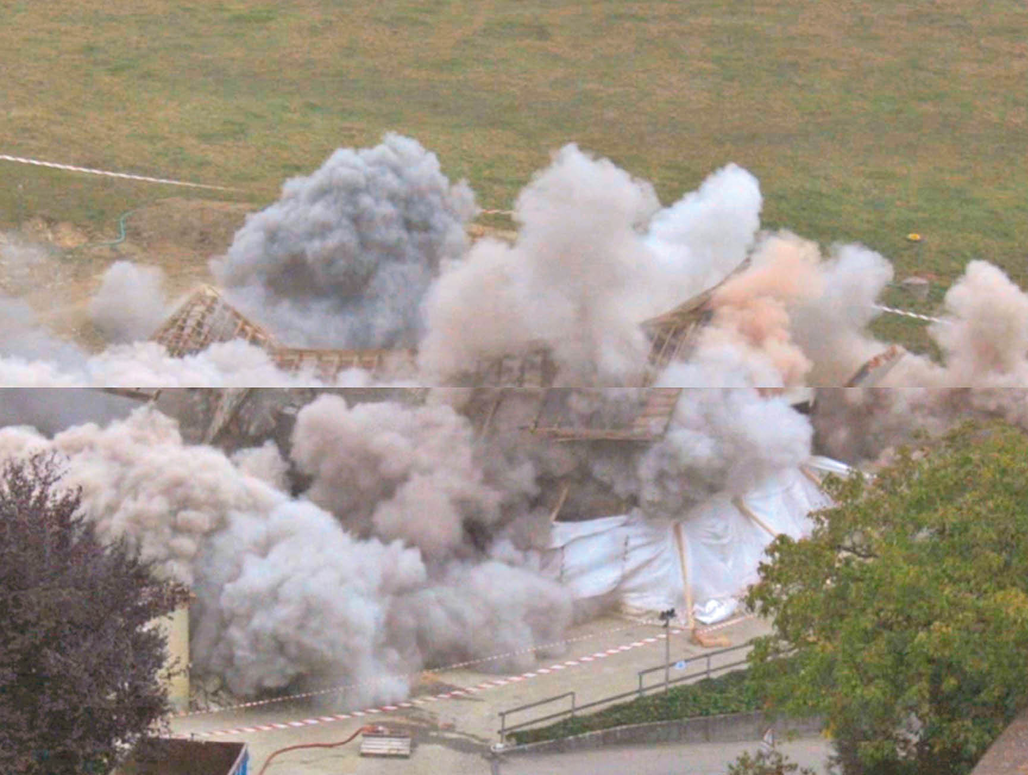 The image is a film still showing the moment of a collapsing building. Big dust clouds erupt around the exploding roof, beams, walls. The middle one can see a line splitting the image in two parts, which results from a time delay introduced by editing the two parts in different running speeds.