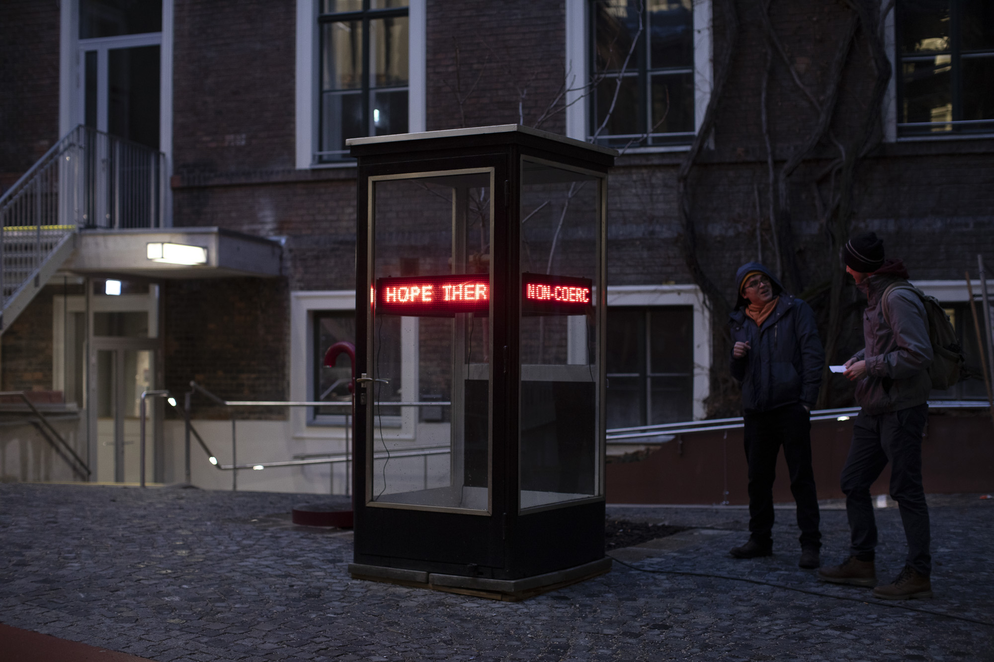 Telephone booth in the WUK courtyard with four LED tickers  installed in it, showing looped red text fragments. Two of the LED tickers face the camera; one of them shows Jenny Holzer’s quotes, and the other shows fragments of Joonas Lahtinen’s letter to Jenny Holzer. Two persons talk to each other next to the telephone booth. Photo.