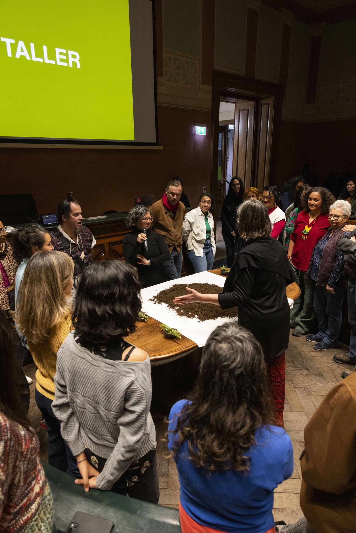 A group of approximately 30 people is standing around a table in the Anatomy Hall of the Academy of Fine Arts Vienna. One person is holding a microphone and one person is standing close to the table and gesturing.