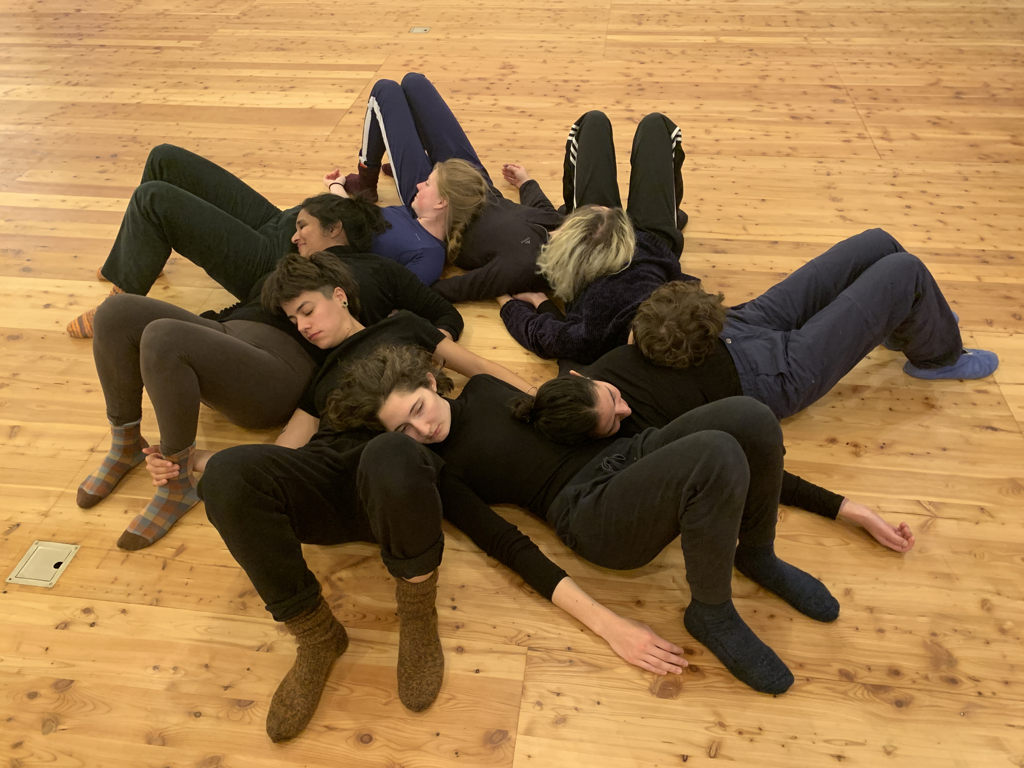Seven people lie interconnected on a wooden floor in various positions, with their bodies overlapping and touching in a circular formation. They are wearing casual dark clothing and appear relaxed with eyes closed, demonstrating a collective embodied practice or group meditation exercise.
