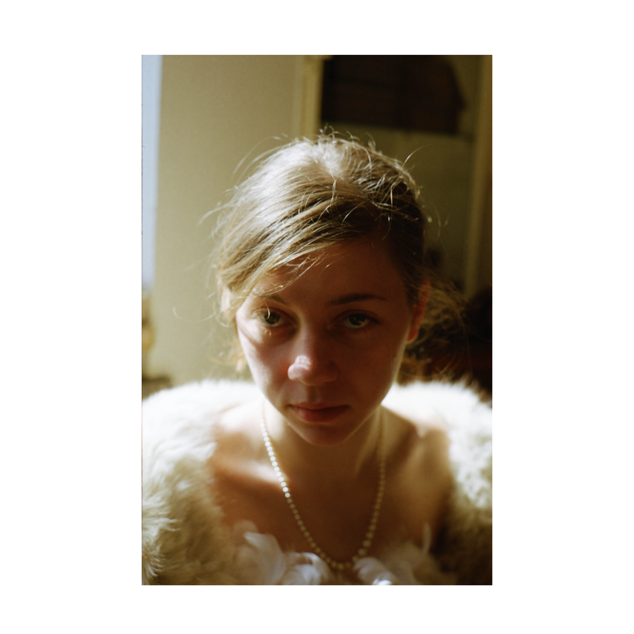 A close-up portrait of a young woman indoors, lit by natural light from the left side. She has light brown hair loosely tied back, with a few strands falling across her forehead. She wears a pearl necklace and a garment trimmed with light fur. The background is softly out of focus, highlighting her face in the frame.