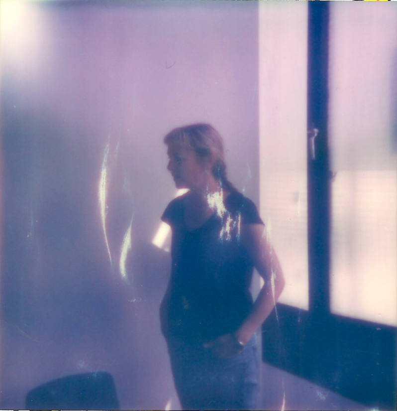 A polaroid photo of a woman wearing a t-shirt standing by a window.