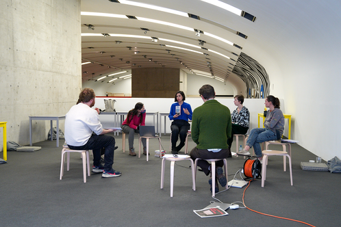 Seven people sit in a circle, the artist Ana de Almeida talks and holds a book in her hand.