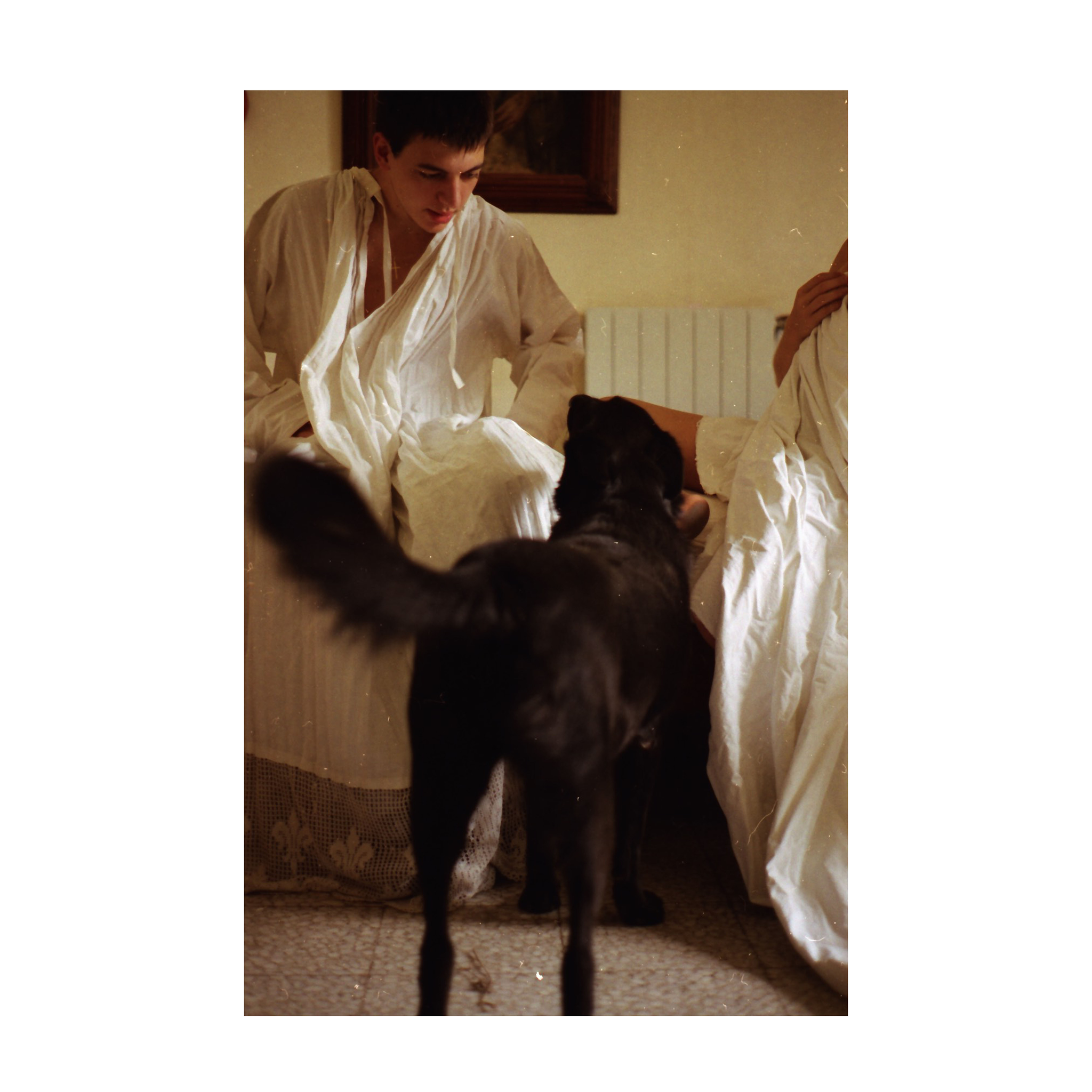 A young man wearing a pleated alb sits on a bed, partially leaning forward. In front of him stands a black dog, seen from behind, its tail raised and mid-motion. Another person, mostly out of frame, is seated beside him, also draped in white fabric. The room has a radiator on the wall, a dark framed picture of Saint Rita above it, and a tiled floor with a lace-trimmed bedcover visible at the bottom of the image.