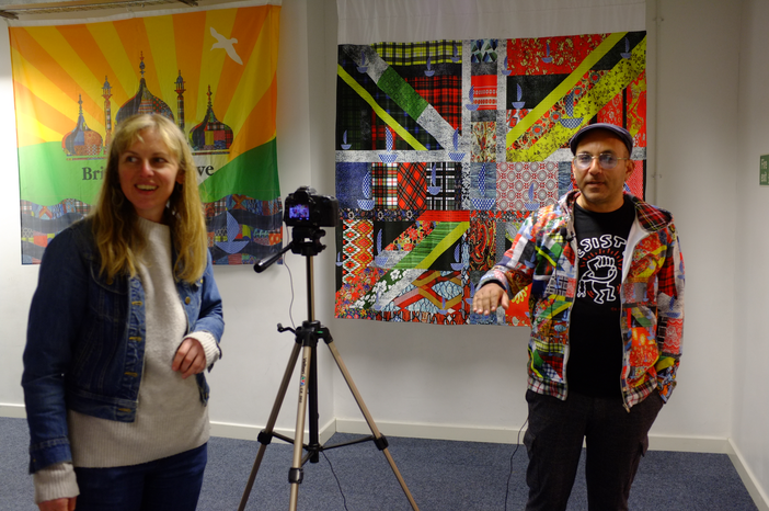 A photo with a woman standing on the left in front of a camera and a man standing on the right in front of a collaged flag.