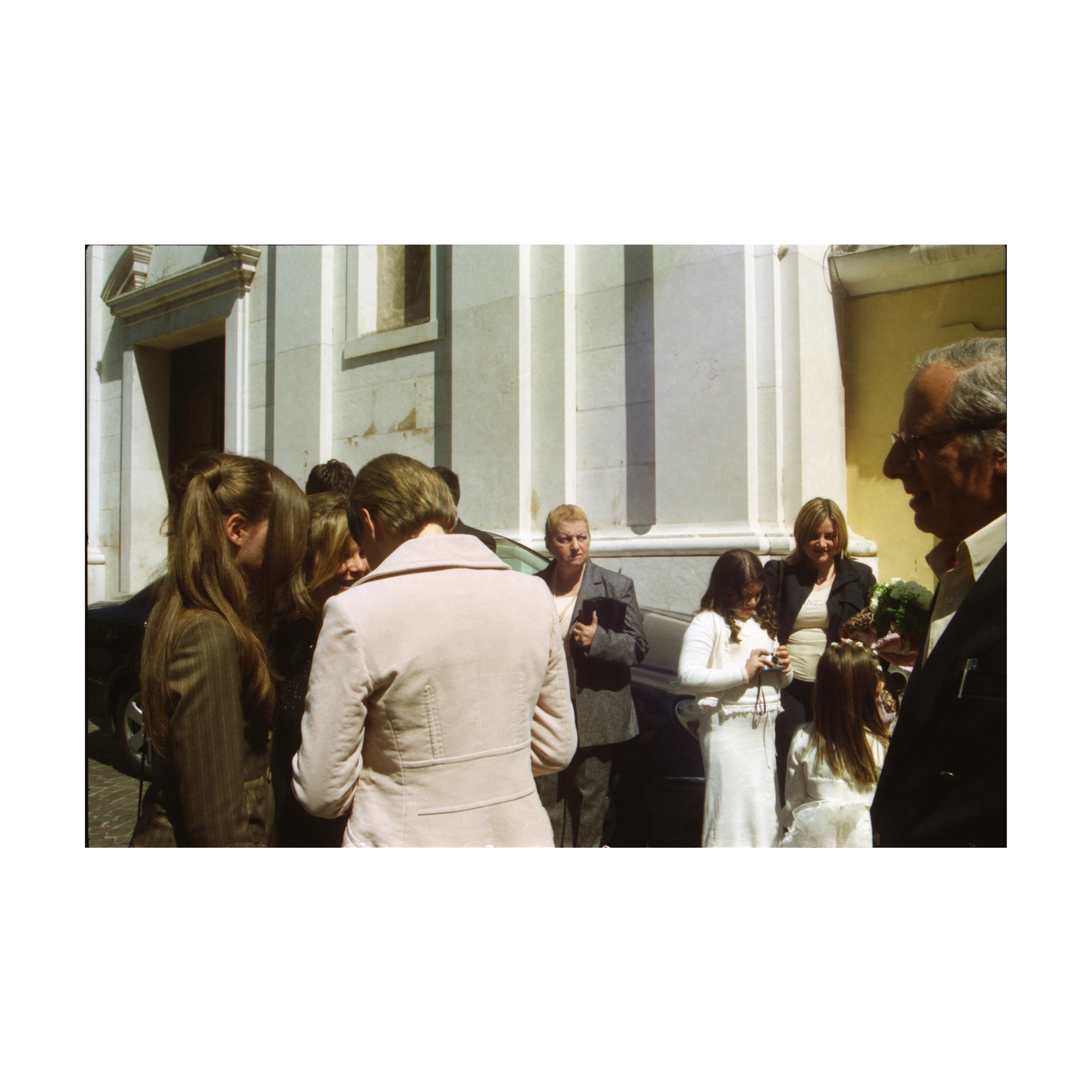 A group of people standing outside a church. Several individuals are dressed in suits and jackets, while two women and a child in white clothing stand near the right side. One man in glasses is in the foreground on the right.