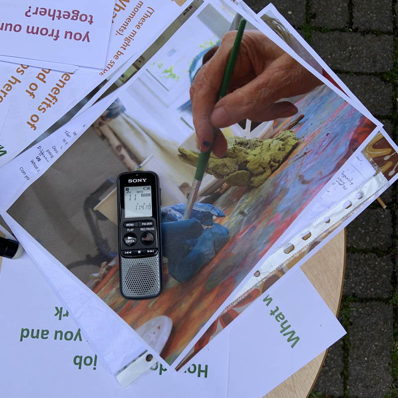 A birds-eye photo of print outs of words and images laid out on a table. The topmost image is of a hand holding a paintbrush painting a clay blue sculpture. On top of the image is an audio recorder