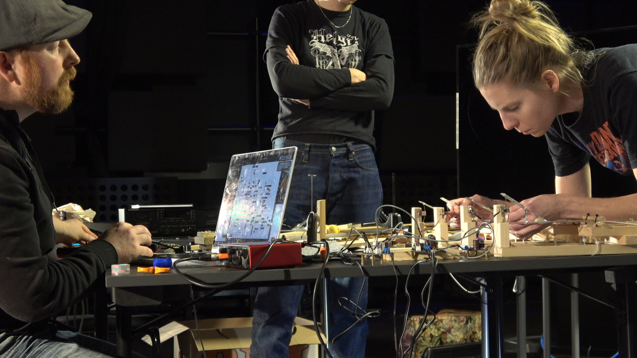 two people at a table across from one another. one person sitting at a laptop and the other standing, playing a wooden, electronic instrument with cassette tape. the three entities are musicking