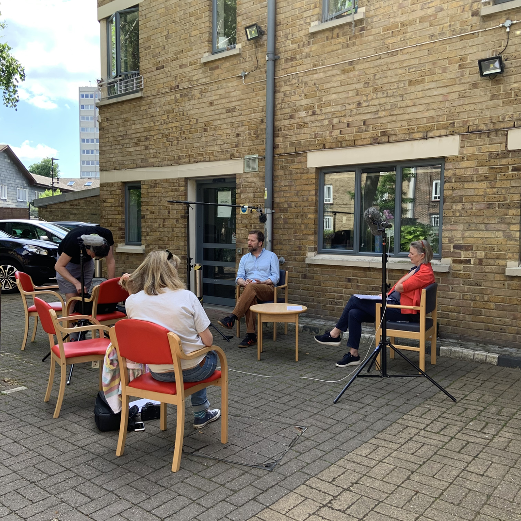 Four people outside the yellow brick building of Mildmay care facility. Some are sitting with microphones and recording equipment nearby
