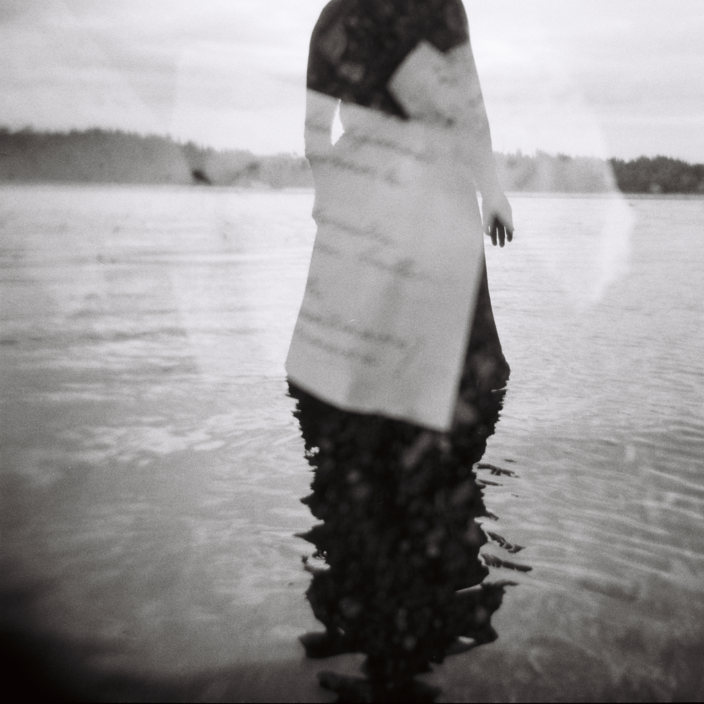 Vedessä seisoo mustaan pitkään hameeseen pukeutunut hahmo, jonka päälle on kaksoisvalotettu runo, jonka tekstiä on vaikea lukea.