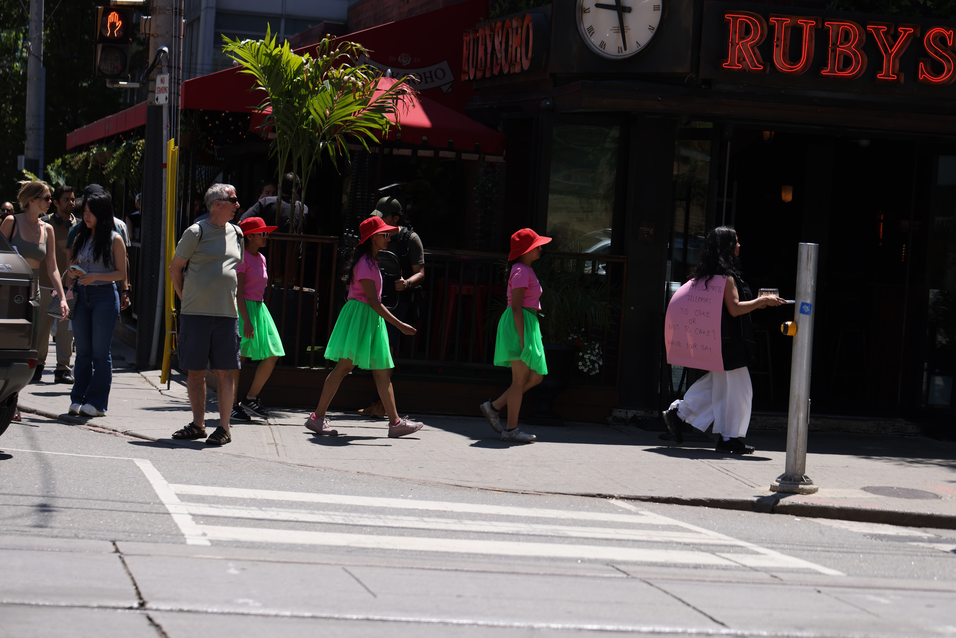 A walk with a playful question, 'To cake or not to cake?', invited pedestrians and patio patrons at the popular destination of King and Portland to reflect within a space largely defined by consumerism, in response to Score 4 created by Mushtari Afroz.
The walk was led by Mushtari, with the question displayed on a piece of pink art paper hanging from her back as she strolled along the sidewalk, carrying a piece of cake on a tray in her hand.
