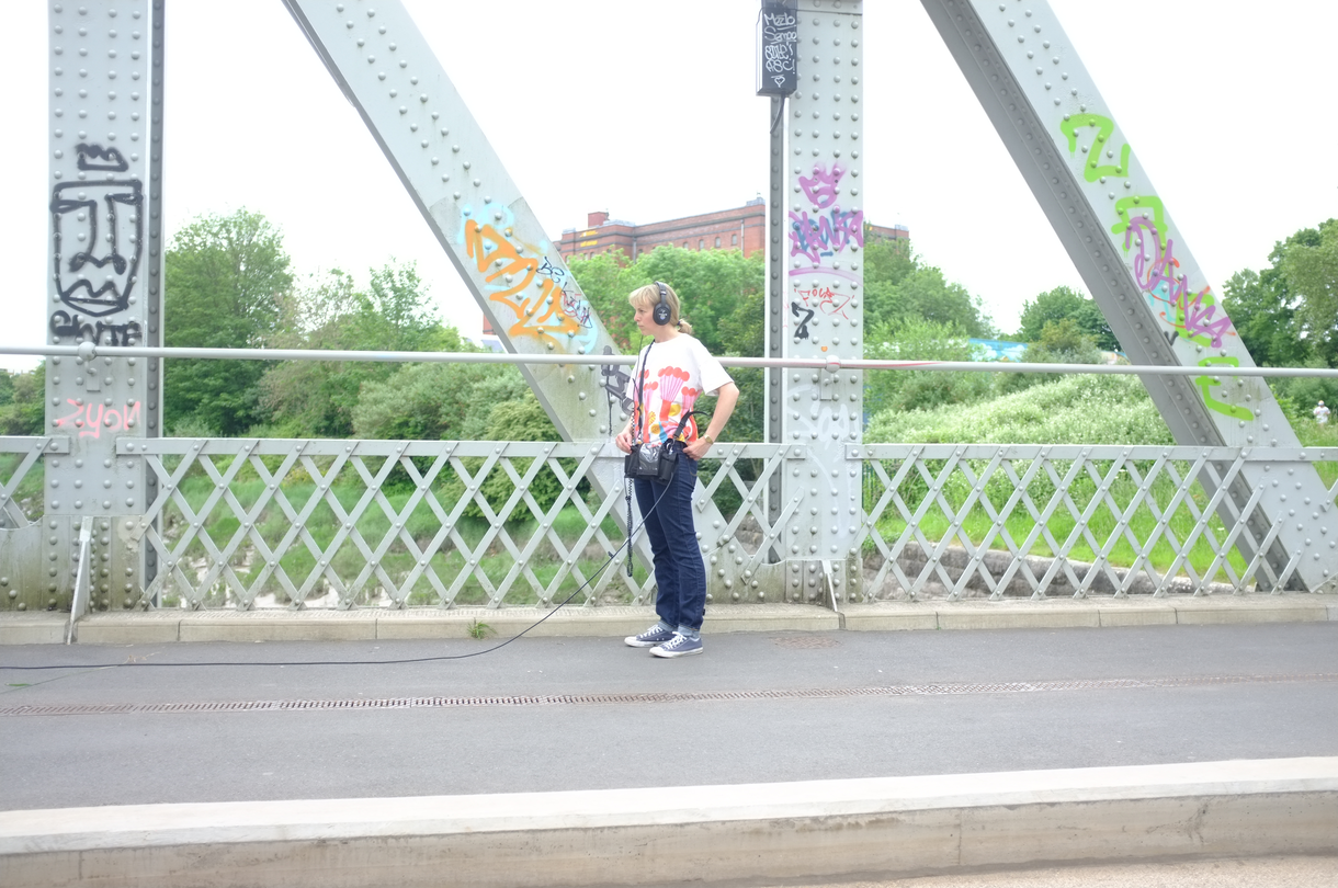 A woman with blonde hair standing on a wrought iron bridge pictured listening to something on headphones. A wire from the headphones reaches to the left of the screen