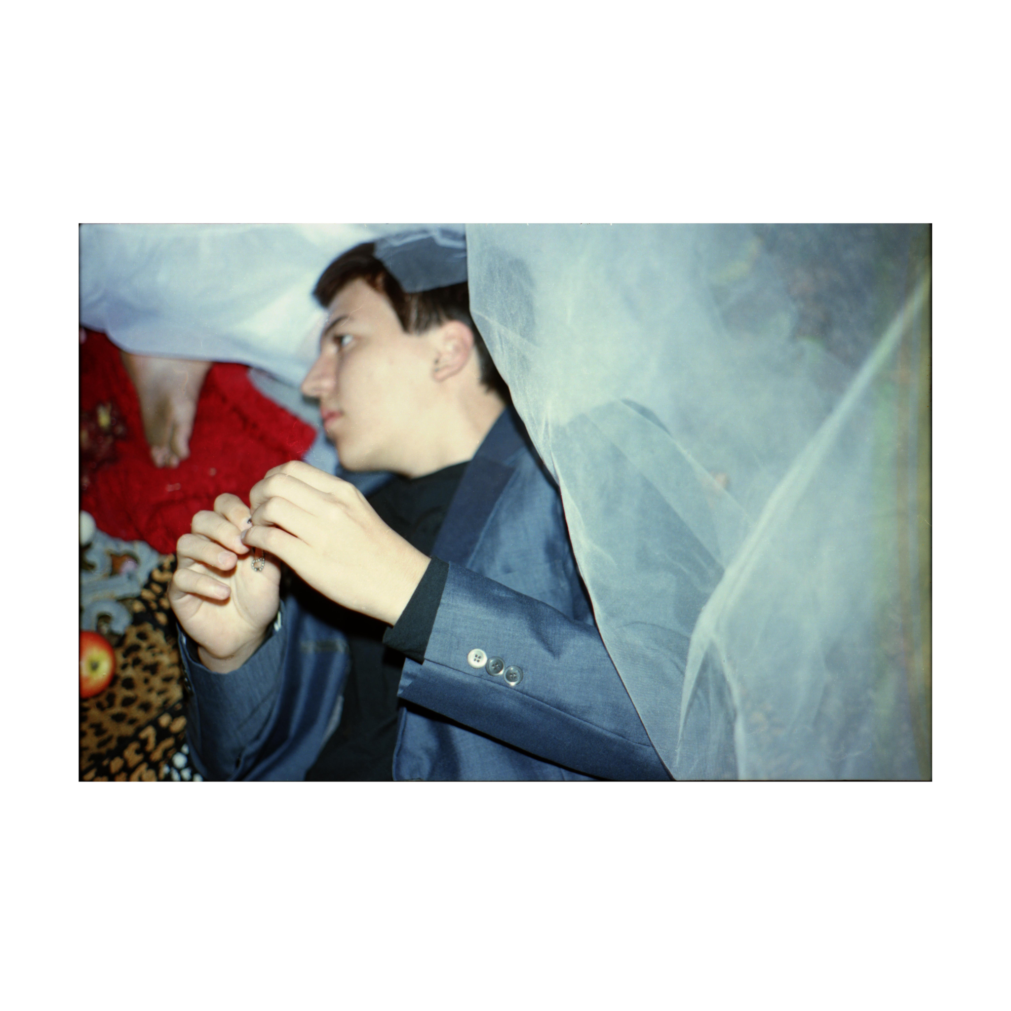 A person in a blue suit jacket lies on patterned fabric, partly covered by a sheer veil. One hand is raised near their face, and the dirty foot of another figure nearby is visible in the frame.